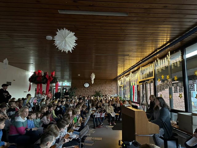 Adventssingen in der Schule: viele Kinder sitzen mit Gitarren zusammen, während eine Erwachsene Klavier spielt.