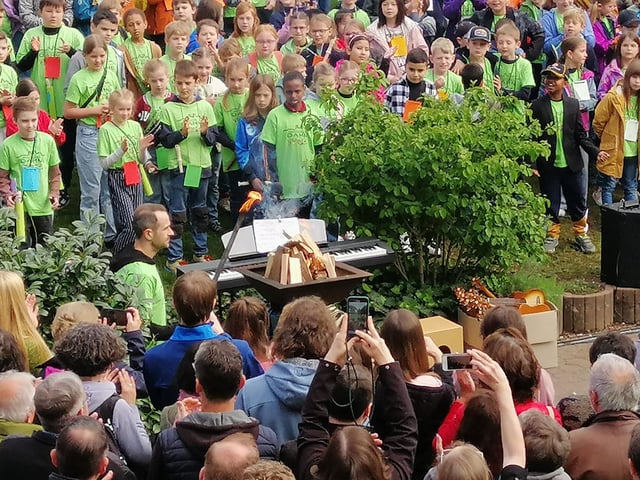 Schulfest im Freien: Kinder in grünen T-Shirts umringen eine Feuerschale, die ein Erwachsener entzündet, Gäste filmen.