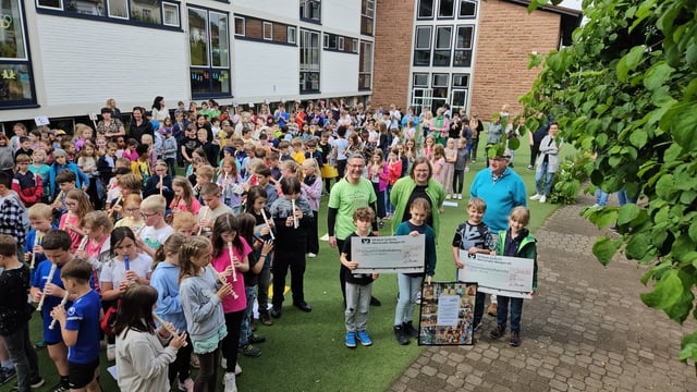 Spendenübergabe auf dem Schulhof: Kinder spielen Blockflöte, während Erwachsene und Kinder große Spendenschecks halten.