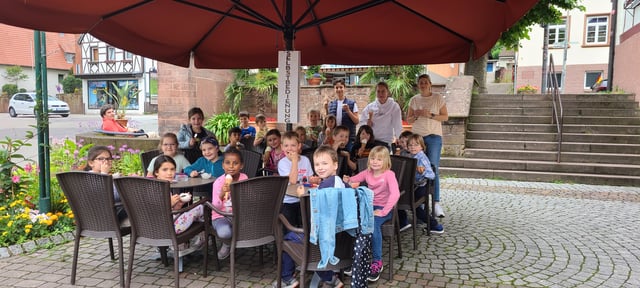 Kinder sitzen beim Ausflug an Tischen unter einem großen Sonnenschirm und essen Eis.