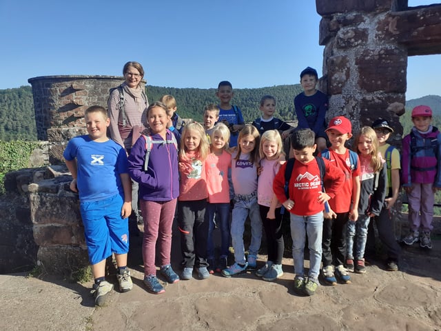 Kinder stehen mit einer Erwachsenen beim Ausflug auf einer Burgruine aus rotem Sandstein, dahinter bewaldete Hügel.
