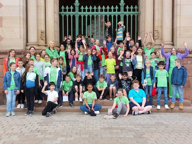 Schülerinnen und Schüler in grünen T-Shirts winken auf den Stufen vor dem verzierten Portal eines historischen Gebäudes.