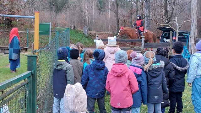 Kinder in Winterjacken beobachten eine rot kostümierte Person, die auf einem braunen Pferd hinter einem Zaun reitet.